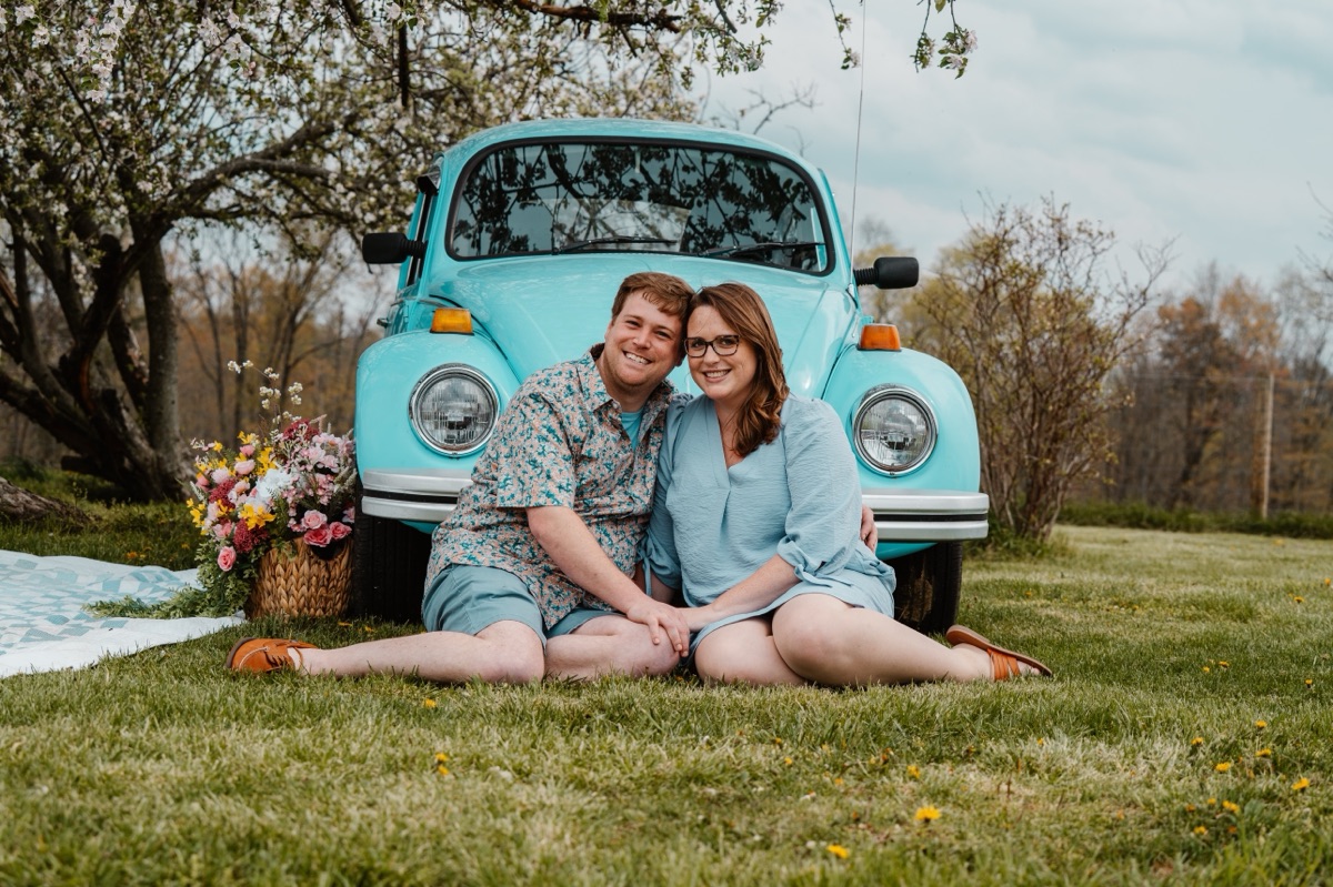 Derek Boyer and his wife Jordan sitting on the grass in front of a turquoise vintage Volkswagen Beetle under a blossoming tree